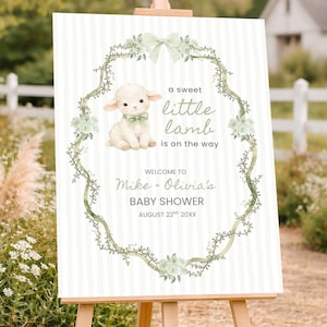 May include: A baby shower sign on a wooden easel. The sign features a watercolor illustration of a lamb with a green bow, surrounded by a floral wreath. Text reads "a sweet little lamb is on the way" and "Mike + Olivia's BABY SHOWER".