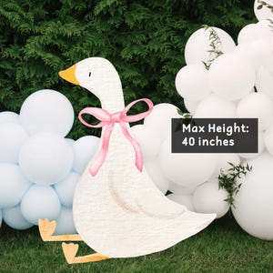 May include: A white goose decoration with a pink bow, standing on green grass, with a backdrop of white and light blue balloons. The goose is 40 inches tall.