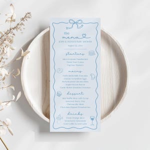 May include: A pale blue baby shower menu with a bow and heart design. The menu lists starters, mains, dessert, and drinks, including mini croissant sandwiches, pancakes, and strawberry lemonade. The menu is on a wooden plate.