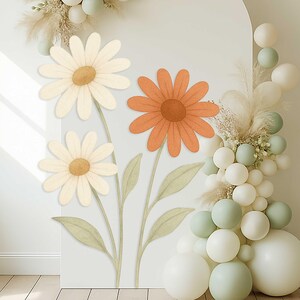 May include: Three paper flowers on a white wall. The flowers are a white daisy, a white daisy, and a peach-colored daisy. The flowers are arranged in a bouquet with green stems and leaves.