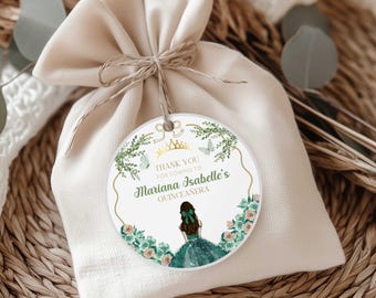 May include: A beige drawstring bag with a round tag. The tag features a gold crown, butterflies, and floral accents. Text on the tag reads "Thank you for coming to Mariana Isabelle's Quinceanera". The illustration shows a girl in a green dress.