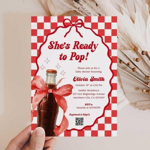 以下が含まれることがあります： 赤と白のチェック柄の背景のベビーシャワーの招待状。招待状には、赤いリボンが付いたソーダボトルの水彩イラストが描かれています。「She's Ready to Pop!」というテキストとイベントの詳細が含まれています。