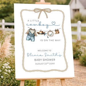 May include: A white sign on a wooden easel with a rope border and the text "A Little Cowboy Is On The Way." The sign welcomes guests to Olivia Smith's baby shower on August 22nd. The sign has illustrations of cowboy-themed items.