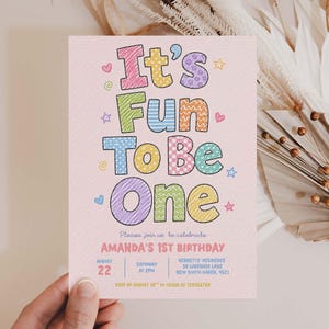 May include: A rectangular invitation card on light pink paper featuring the colourful text It's Fun To Be One in various patterns. Below, it reads Please join us to celebrate AMANDA'S 1ST BIRTHDAY, with date, time, location, and RSVP details included.