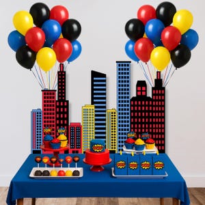 May include: A party scene with a blue tablecloth, cityscape backdrop, and colourful balloons. The table displays cupcakes, cake pops, a cake, and gift bags, all decorated with comic book-style graphics.
