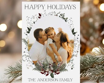 May include: A holiday greeting card with the text "HAPPY HOLIDAYS" and "THE ANDERSON FAMILY". The card features a family photo surrounded by a floral wreath with green and burgundy accents. The family is kissing the child.