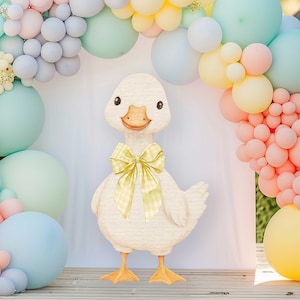 Puede incluir: Una decoración de ganso blanco con un lazo de cuadros amarillos, sobre un fondo de globos de colores pastel. El ganso tiene patas naranjas y un cuerpo texturizado, ideal para un baby shower o fiesta.