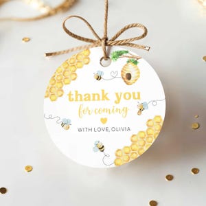 May include: A round white gift tag with a bee theme. The tag features yellow text that reads "thank you for coming" and "with love, Olivia". The tag has illustrations of bees, a beehive, and honeycomb.
