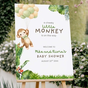 May include: A baby shower sign on a wooden easel. The sign features a cartoon monkey holding balloons, with text that reads "a cheeky little MONKEY is on the way." The sign also includes the names "Mike and Olivia" and the date.