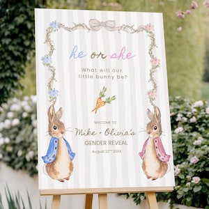 May include: A gender reveal sign on a wooden easel. The sign features a watercolor illustration of two bunnies, one in a blue jacket and one in a pink jacket. Text includes "He or She" and "What will our little bunny be?"
