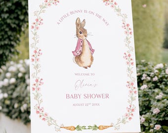 May include: A white sign on a wooden easel with the text "A LITTLE BUNNY IS ON THE WAY" and "WELCOME TO Olivia's BABY SHOWER AUGUST 22 20XX". The sign features a watercolor illustration of a bunny wearing a pink jacket, surrounded by a floral and carrot border.