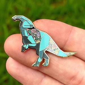 Op de afbeelding: Een turquoise en grijze emaille pin in de vorm van een dinosaurus met een bloemenpatroon op de rug. De pin heeft een gouden omtrek en wordt in een hand gehouden tegen een vage groene achtergrond.
