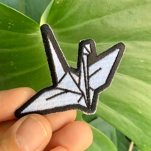 May include: White origami crane iron-on patch with black stitching. The patch is held in a hand against a green leafy background.