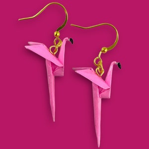May include: A pair of pink origami flamingo earrings with gold hooks. The earrings are made of paper and are a delicate and whimsical accessory.