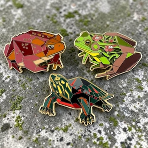 May include: Three geometric animal pins: a red-brown toad, a green frog with floral accents, and a turtle with red and green details. Each pin has a gold outline and a unique polygonal design.