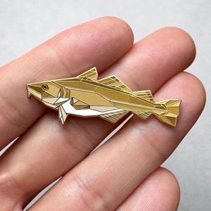 Könnte beinhalten: Ein goldfarbener geometrischer Fisch-Ansteckpin mit detailliertem Design. Der Pin zeigt eine stilisierte Darstellung eines Fisches mit scharfen Winkeln und Facetten. Der Fisch hat einen weißen Bereich am Bauch und wird in einer Hand gehalten.