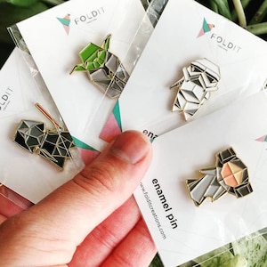 May include: A hand holding four enamel pins in clear packaging. The pins feature geometric designs of characters in gold trim. The packaging has the text "FOLDIT" and "enamel pin" with a website address.
