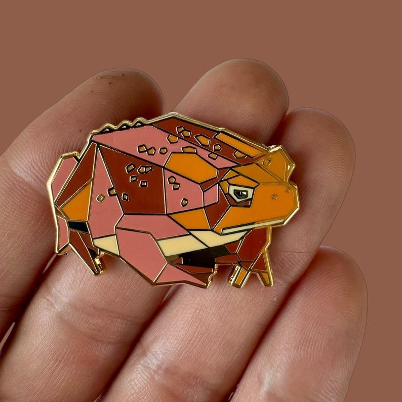 May include: A geometric enamel pin featuring a stylized frog design. The pin showcases a color palette of pink, orange, and brown, with gold-colored metal accents. The frog's form is segmented into polygonal shapes, giving it a modern, artistic look.