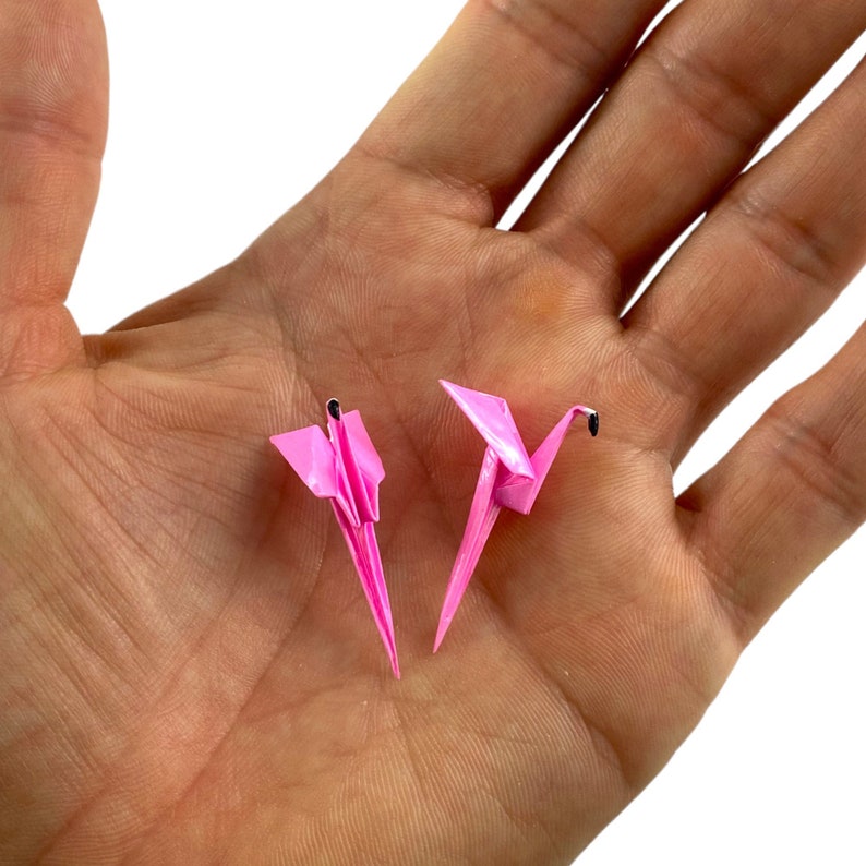 May include: Two pink origami cranes, folded from paper, are displayed on an open hand.