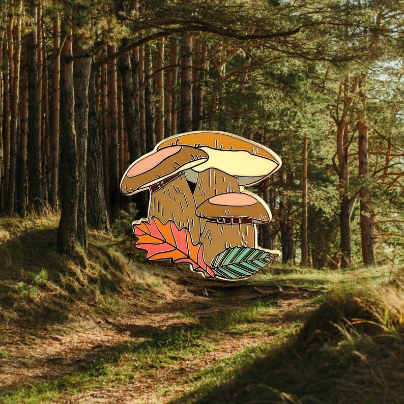 Pu&ograve; includere: Uno spillo in smalto dorato con tre funghi con cappelli e gambi marroni e beige. I funghi sono circondati da un gruppo di foglie autunnali in tonalit&agrave; di arancio, giallo e verde.