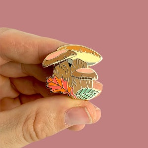 Pu&ograve; includere: Spilla in smalto dorata con un gruppo di tre funghi con accenti marroni, arancioni e gialli. I funghi sono circondati da una foglia verde e arancione.