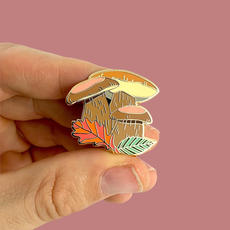 Pu&ograve; includere: Spilla in smalto dorata con un gruppo di tre funghi con accenti marroni, arancioni e gialli. I funghi sono circondati da una foglia verde e arancione.