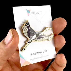 Puede incluir: Pin de esmalte dorado de una gr&uacute;a origami geom&eacute;trica en vuelo. El pin est&aacute; en una tarjeta blanca con el texto "FOLDIT CREATIONS" y "enamel pin" y la direcci&oacute;n del sitio web "www.folditcreations.com".