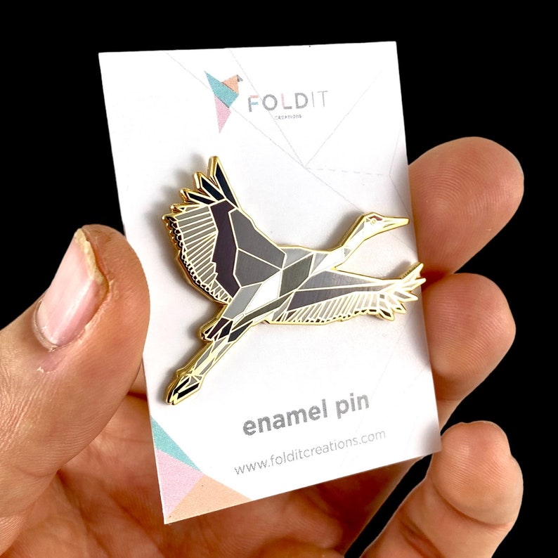 Puede incluir: Pin de esmalte dorado de una gr&uacute;a origami geom&eacute;trica en vuelo. El pin est&aacute; en una tarjeta blanca con el texto "FOLDIT CREATIONS" y "enamel pin" y la direcci&oacute;n del sitio web "www.folditcreations.com".