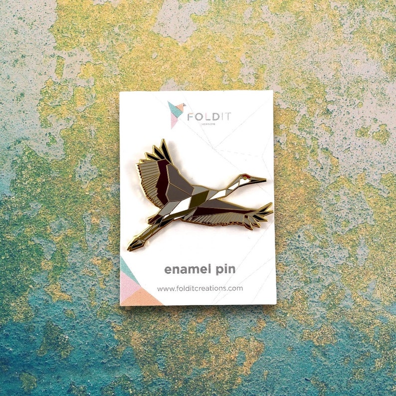 May include: A gold enamel pin featuring a geometric design of a crane in flight. The pin is on a white card with the text "FOLDIT Jewelry" and "enamel pin" printed on it.