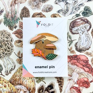 Pu&ograve; includere: Una spilla in smalto dorata con tre funghi marroni con foglie arancioni e verdi. La spilla &egrave; su una carta bianca con il testo "Foldit Creations" e "enamel pin" in nero.