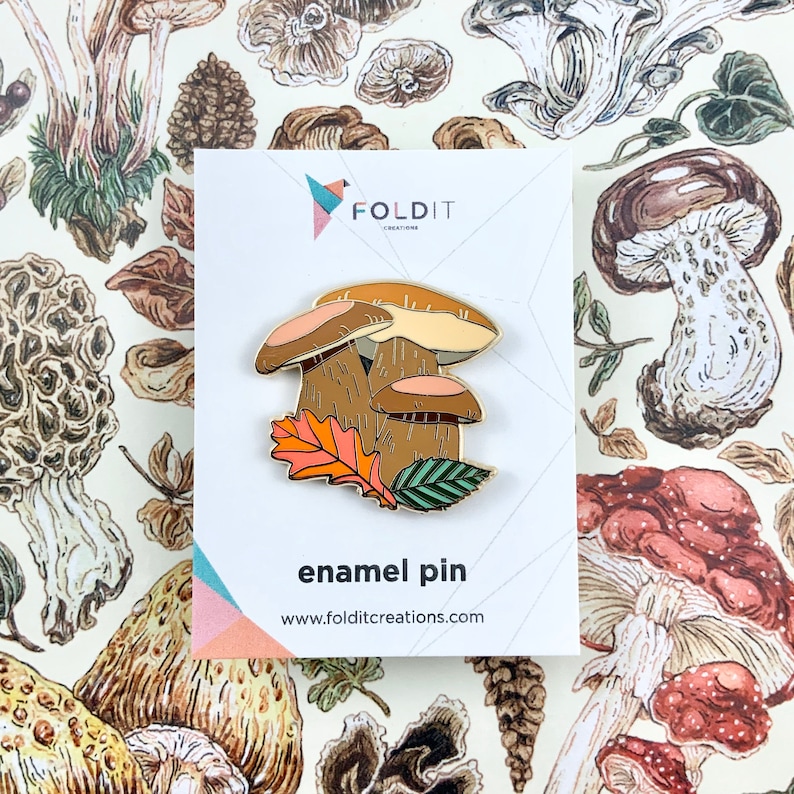 Pu&ograve; includere: Una spilla in smalto dorata con tre funghi marroni con foglie arancioni e verdi. La spilla &egrave; su una carta bianca con il testo "Foldit Creations" e "enamel pin" in nero.