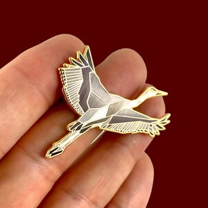 Puede incluir: Un pin de esmalte dorado con un dise&ntilde;o geom&eacute;trico de una gr&uacute;a en vuelo. La gr&uacute;a est&aacute; representada en tonos de gris y blanco con detalles dorados.