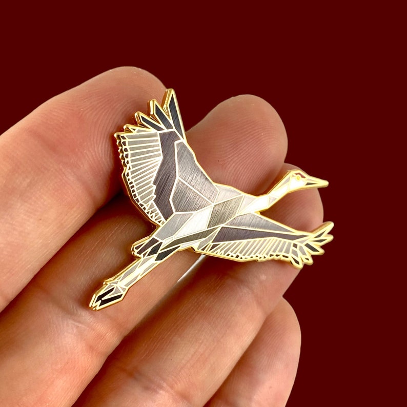 Puede incluir: Un pin de esmalte dorado con un dise&ntilde;o geom&eacute;trico de una gr&uacute;a en vuelo. La gr&uacute;a est&aacute; representada en tonos de gris y blanco con detalles dorados.