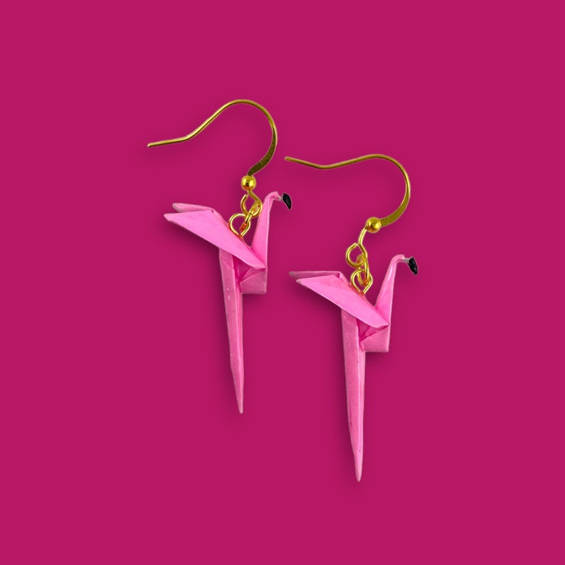 May include: A pair of pink origami flamingo earrings with gold hooks. The earrings are made of paper and are a delicate and whimsical accessory.