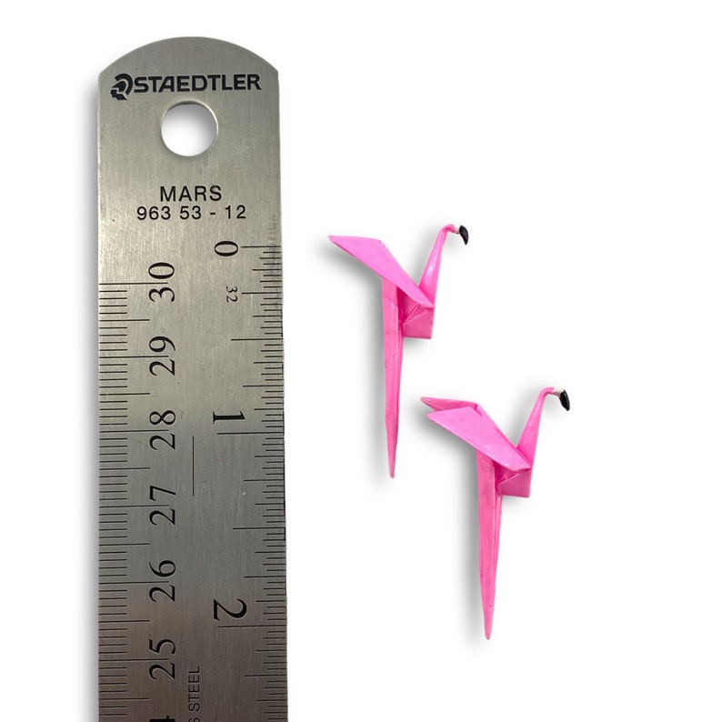 May include: Two pink origami flamingo paper birds, each about 2 inches tall, are shown next to a ruler. The ruler is marked in inches and centimeters.
