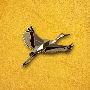 Puede incluir: Un pin de metal de color dorado en forma de gr&uacute;a con patrones geom&eacute;tricos en negro, blanco y gris. La gr&uacute;a est&aacute; en vuelo con las alas extendidas.