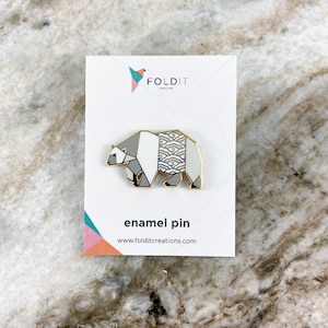 May include: A white and gray enamel pin featuring a geometric origami bear design with a wave pattern on the back. The pin is on a white card with the text "Foldit Creations" and "enamel pin" printed on it.