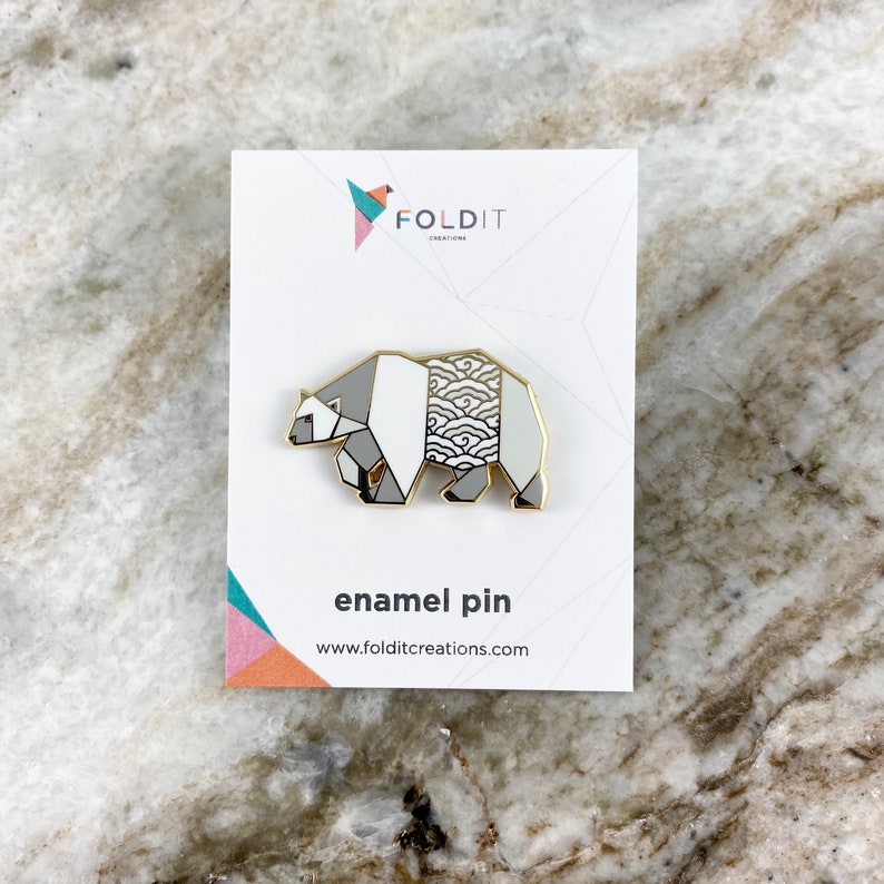 May include: A white and gray enamel pin featuring a geometric origami bear design with a wave pattern on the back. The pin is on a white card with the text "Foldit Creations" and "enamel pin" printed on it.