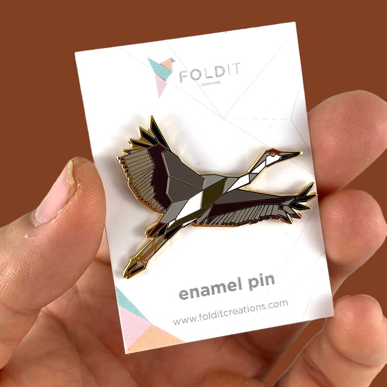 Puede incluir: Un pin de esmalte dorado con una gr&uacute;a geom&eacute;trica en vuelo. La gr&uacute;a est&aacute; compuesta por tri&aacute;ngulos negros, blancos y grises. El pin est&aacute; en una tarjeta blanca con el texto "FOLDIT CREATIONS" y "enamel pin" en negro.