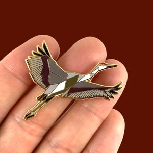 Puede incluir: Un pin de esmalte dorado con un dise&ntilde;o geom&eacute;trico de una gr&uacute;a en vuelo. La gr&uacute;a est&aacute; representada en tonos de gris, blanco y burdeos, con un pico rojo y negro.