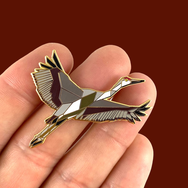 Puede incluir: Un pin de esmalte dorado con un dise&ntilde;o geom&eacute;trico de una gr&uacute;a en vuelo. La gr&uacute;a est&aacute; representada en tonos de gris, blanco y burdeos, con un pico rojo y negro.