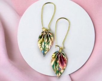 Könnte beinhalten: Ein Paar goldene Ohrringe mit floral gemusterten, blattartigen Anhängern. Die Ohrringe liegen auf einer weißen Oberfläche.