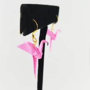 May include: A pair of pink origami crane earrings with gold hooks. The cranes are hanging from a black display stand.