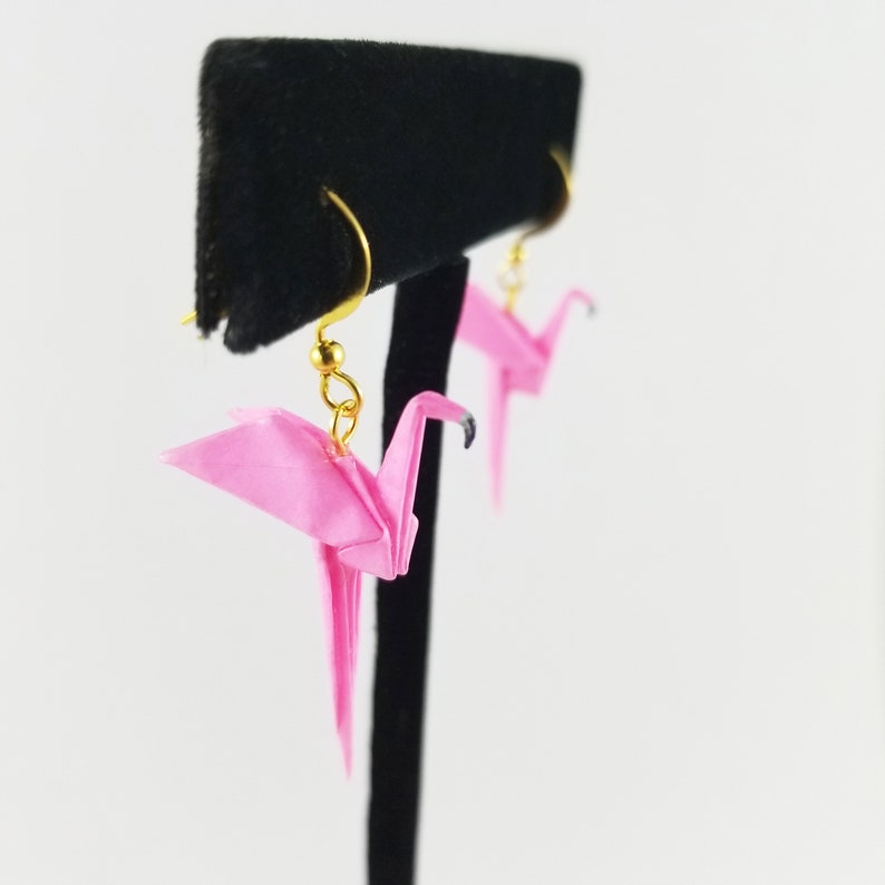 May include: A pair of pink origami crane earrings with gold hooks. The cranes are hanging from a black display stand.