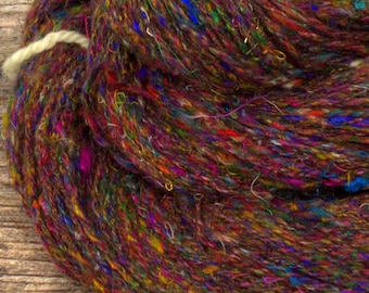 May include: A skein of colorful yarn, primarily brown with flecks of blue, yellow, and red. The yarn is tightly wound and has a textured appearance. The yarn is on a wooden surface.