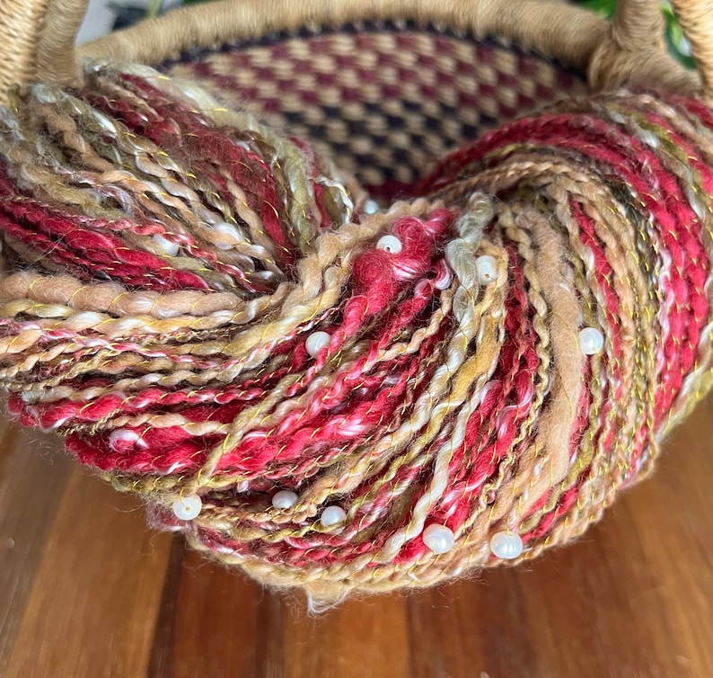 May include: Close-up of a ball of yarn with a mix of red, gold, beige, and white fibres. The yarn is adorned with small, white beads. The basket's woven rim is visible in the background.