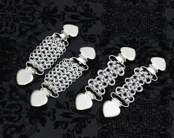 May include: Four silver sweater clips with heart-shaped ends and chainmail-style links. The clips have a shiny finish and are arranged on a dark, patterned background. These decorative clips can be used to fasten cardigans or other garments.