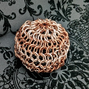 May include: A spherical object constructed from interlocking copper-colored metal rings. The structure features a dense, woven chainmail pattern that creates a textured, rounded form with a metallic sheen and visible gaps between the individual links.