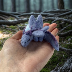 Op de afbeelding: Een klein naaldgevilte drakenfiguurtje in tinten maagdenpalmblauw en lavendel. Het wezen heeft zachte, pluizige texturen, puntige oren, gevouwen vleugels en donker gestikte ogen, vervaardigd uit wolvezels in een compact, handzaam formaat.