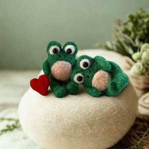 May include: Two green felt frog figurines with large eyes and pink bellies sit on a cream-coloured, rounded surface. A small red heart is placed next to one frog. The frogs are handmade and whimsical.